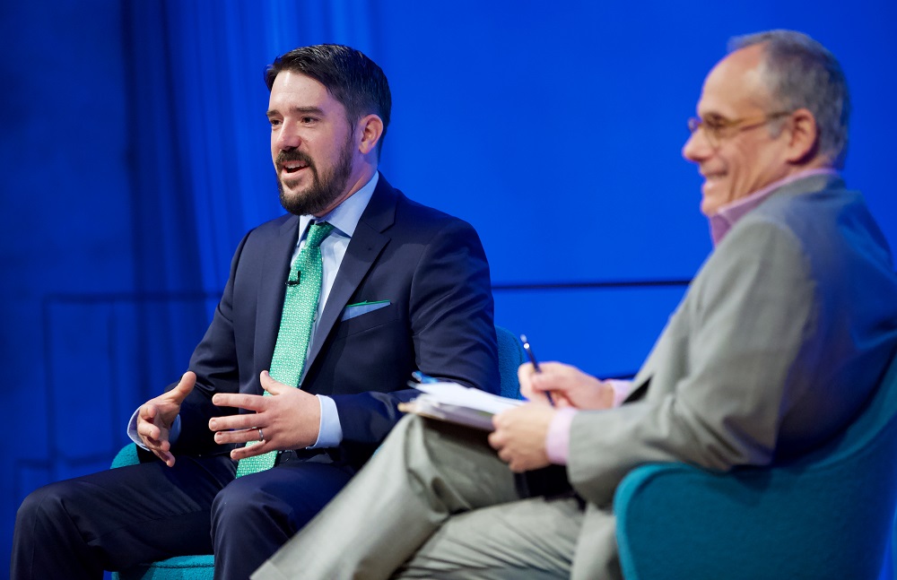 Two men sit on a blue-lit auditorium stage. One addresses an unseen audience while the other smiles.