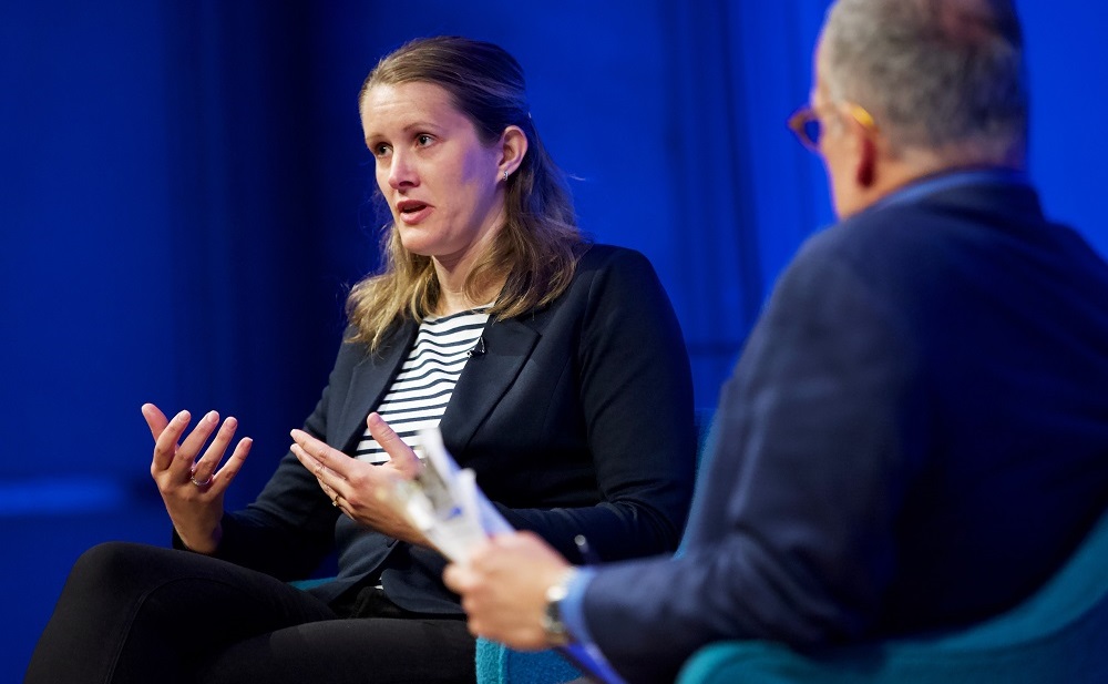 A female speaker gestures with her hands while the male moderator looks on.