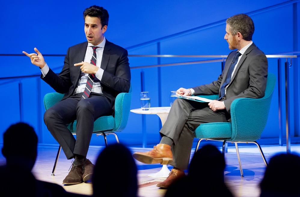 Two men in suits, a public program participant and a moderator, sit on a blue-lit auditorium stage. The program participant gestures with his hands while speaking out to the audience, whose heads appear in silhouette. The moderator looks on.