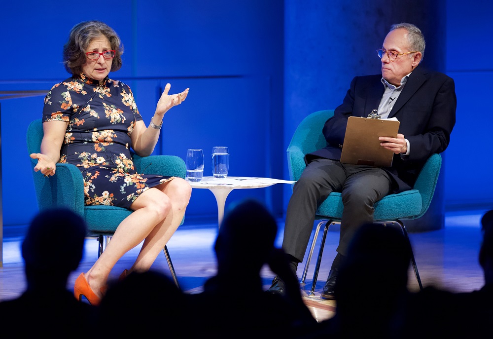 A public program participant sits on a blue-lit auditorium stage and gestures with her hands while looking out into the audience, whose heads appear in the foreground in silhouette. The program moderator sits to her left and looks on.