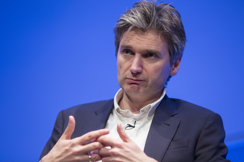 In this close-cropped photograph, a man with salt-and-pepper hair addresses an audience off-camera while gesturing with his hands.