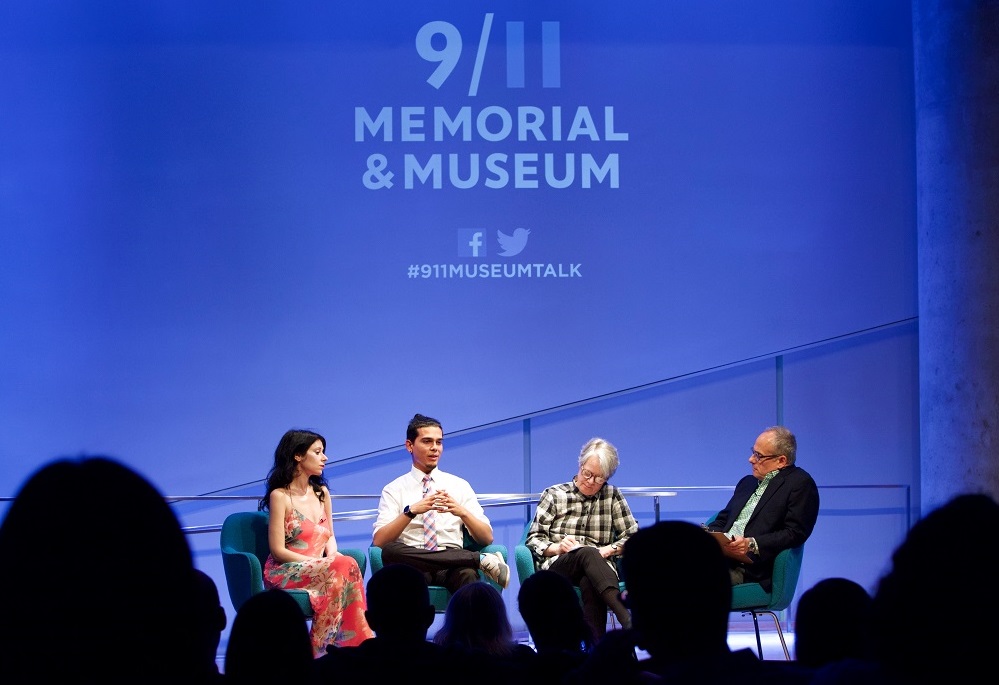 Four people sit on a blue-lit auditorium stage during a public program. The heads of audience members appear in silhouette in the foreground.