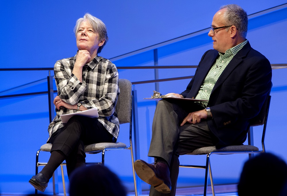 One woman with crossed legs sits on a blue-lit auditorium stage and looks up at the ceiling in thoughtful repose.