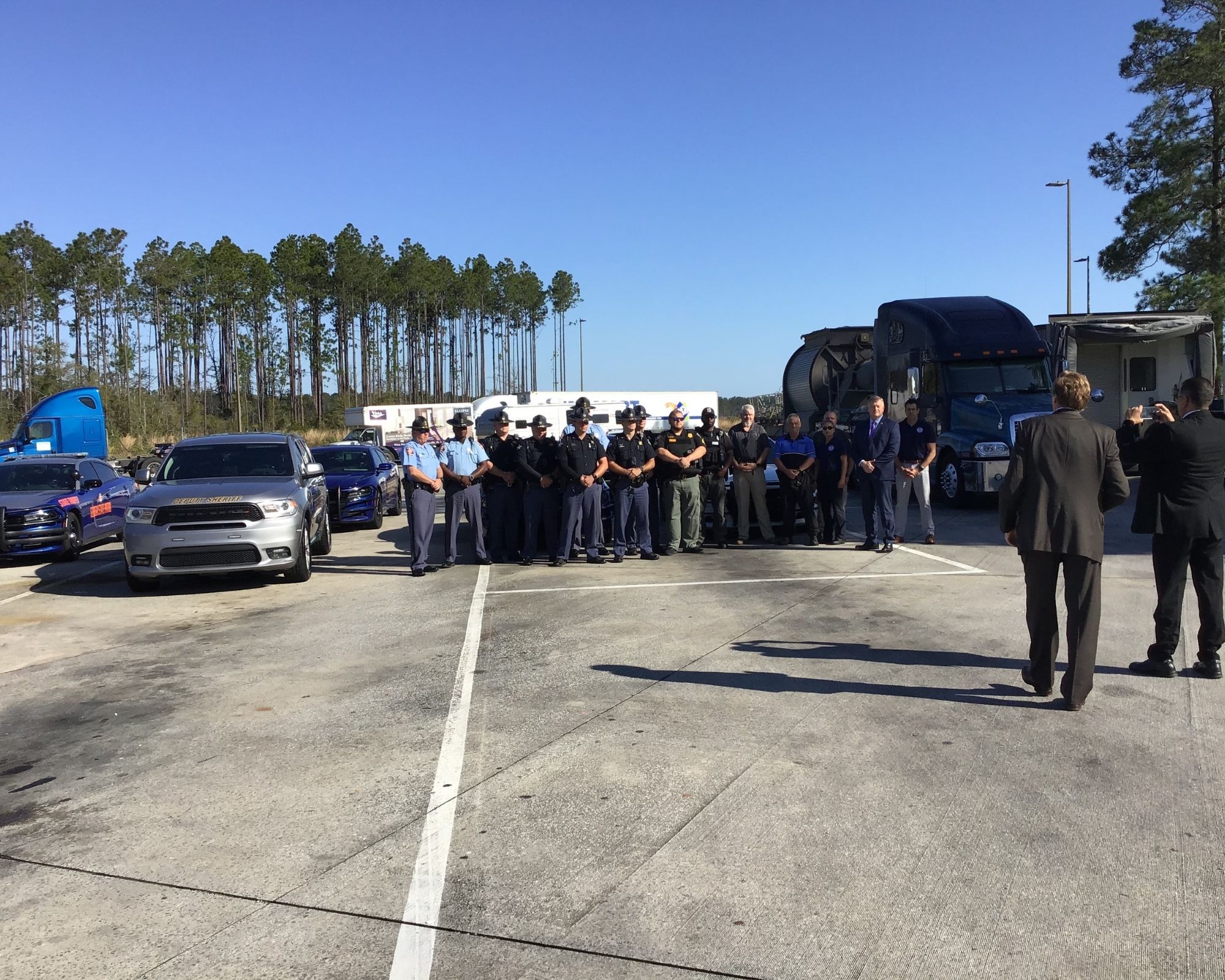 A group of law enforcement and government officials stand against a blue sky with green palm trees