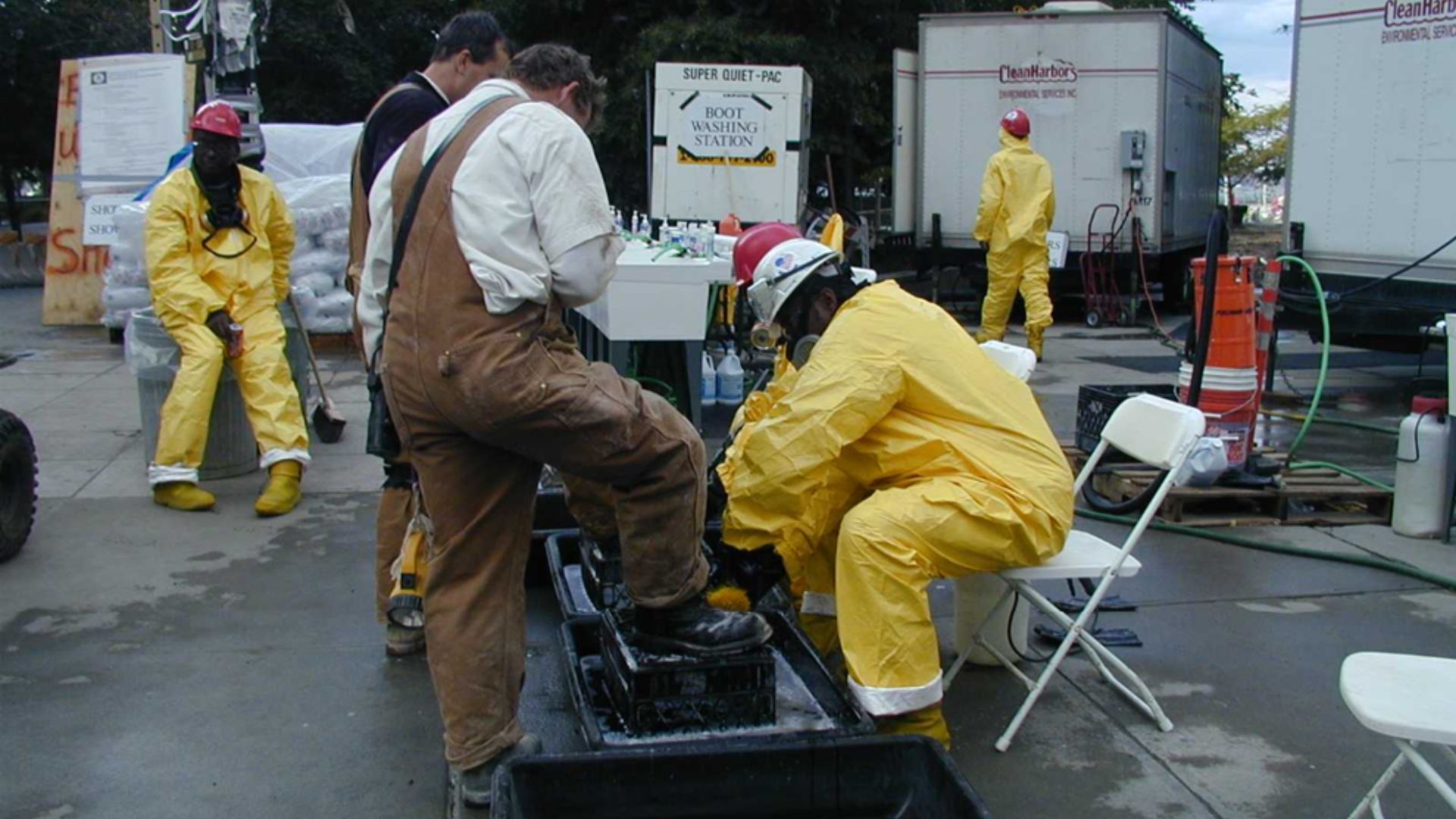 Two workers get their boots washed by two other workers in yellow hazmat uniforms.