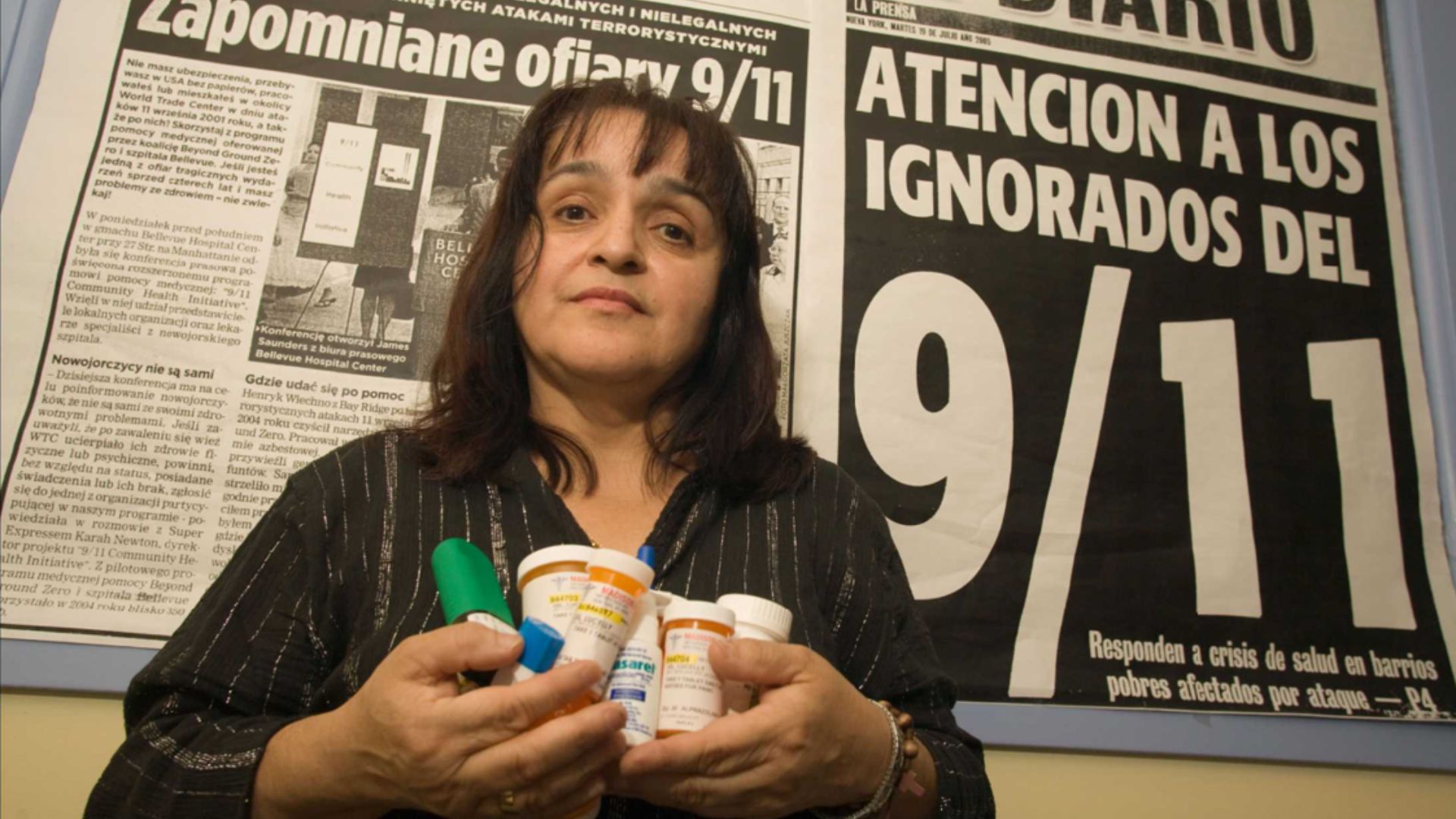 A dark haired woman holds several pill bottles while standing against a wall with 9/11 newspaper clippings.