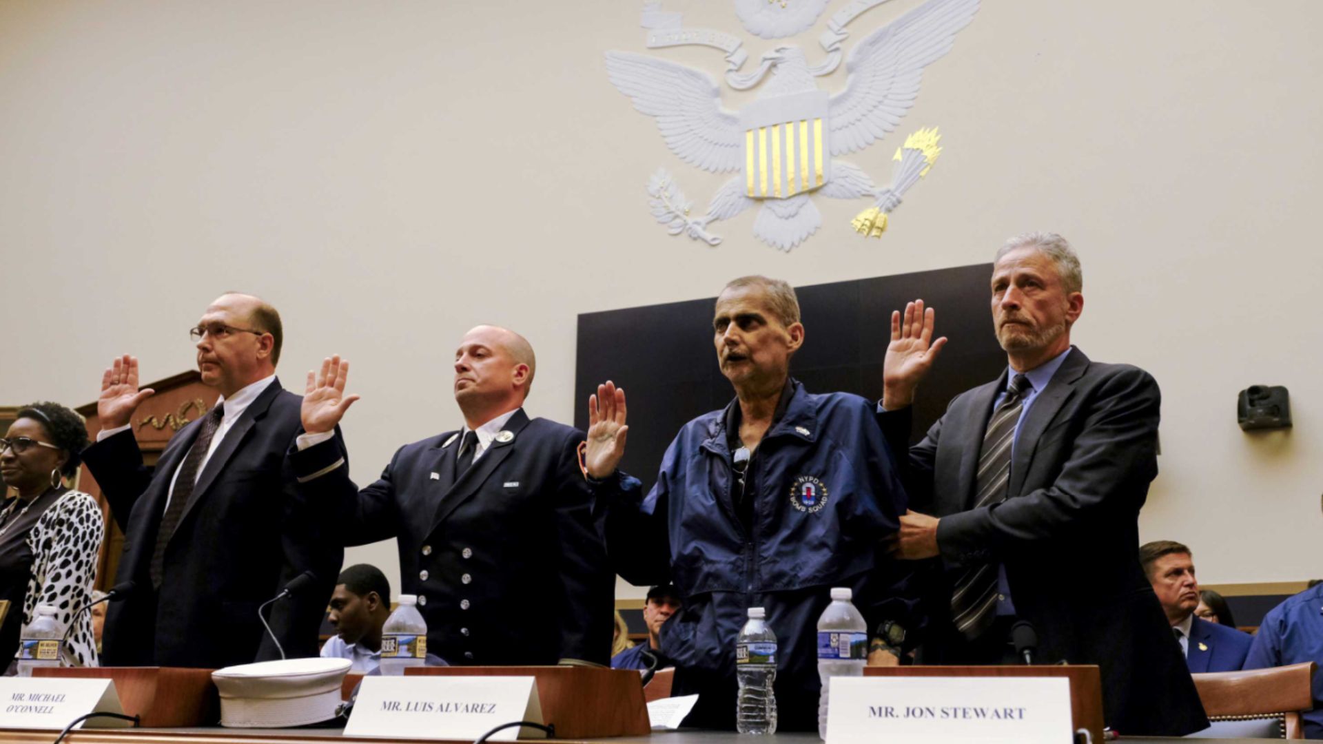 Four people raise their left hands while being sworn in before testifying. The wall behind them is cream-colored.