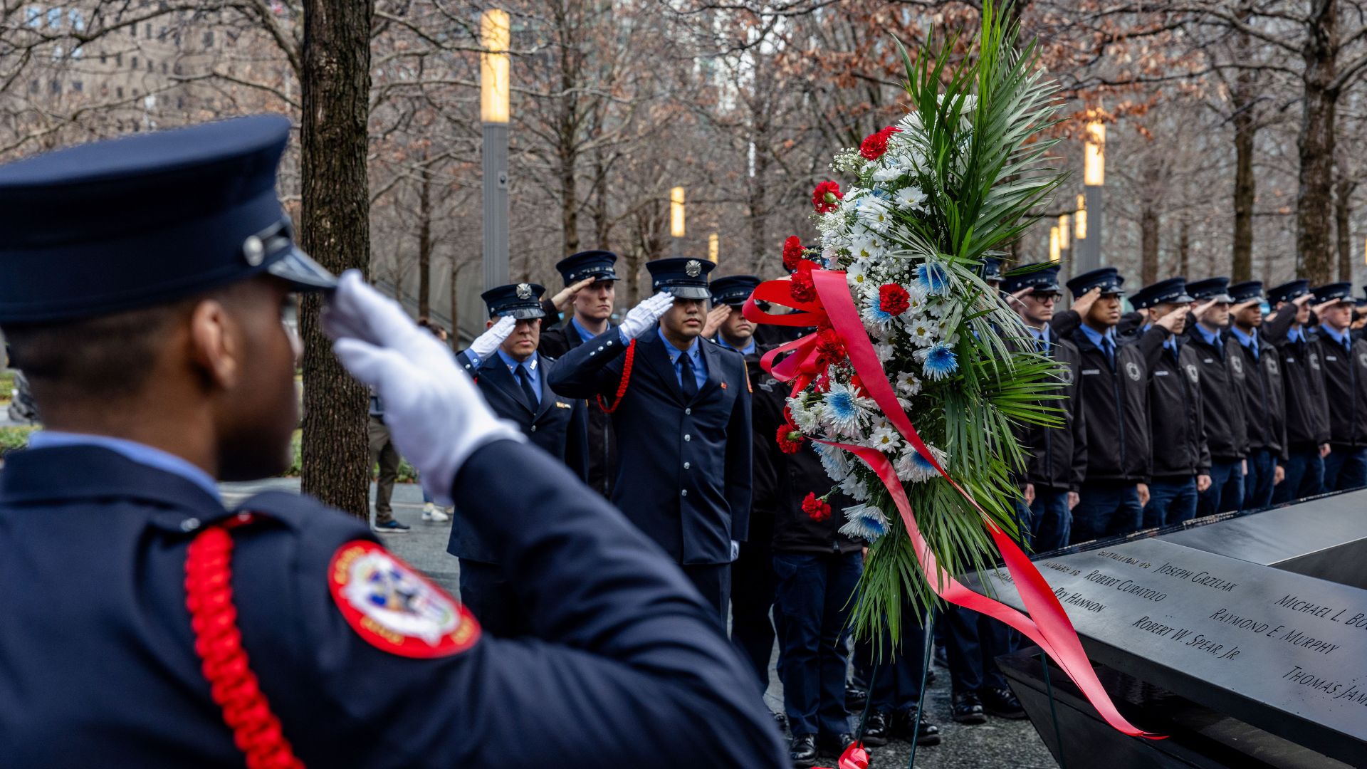 Probationary FDNY officers in uniform along the Memorial. At the foreground, a back view of one probie saluting.