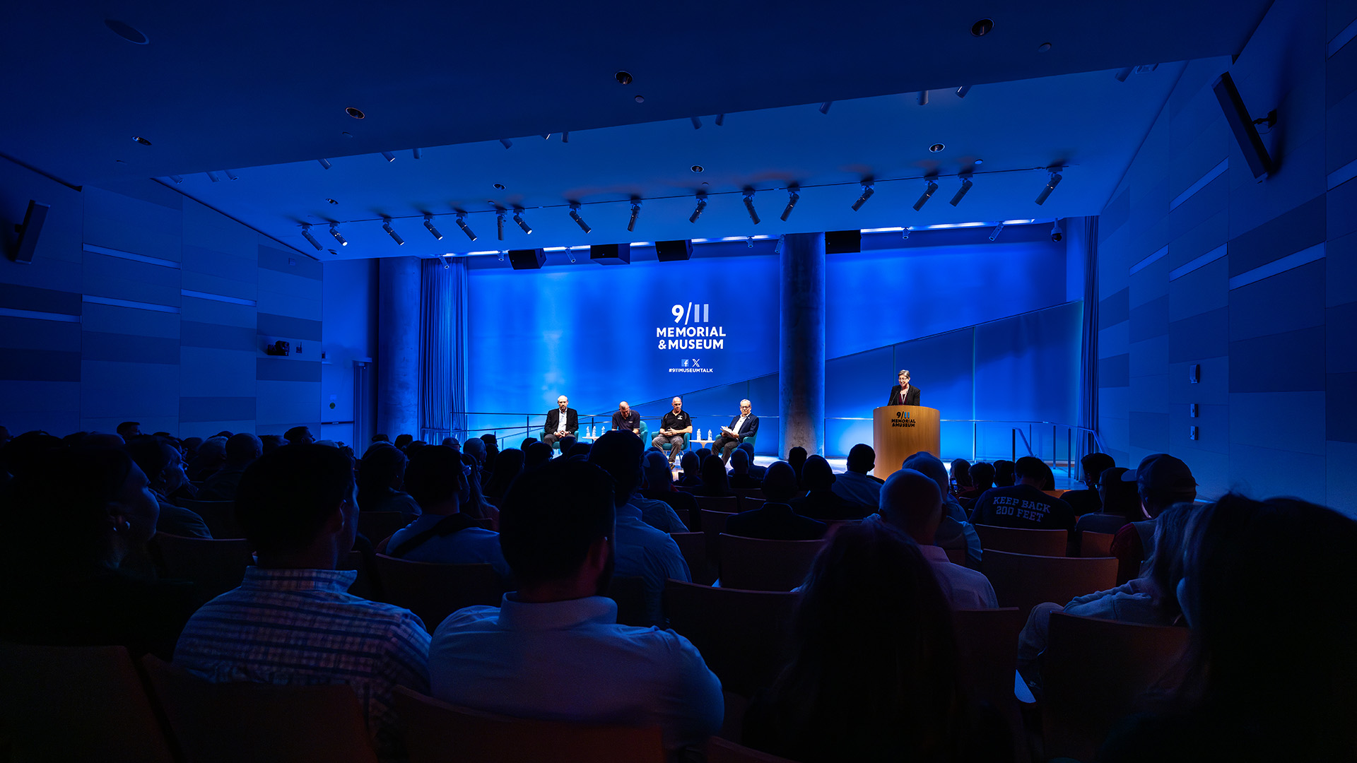 a wide view of the entire 9/11 Memorial and Museum Stage, with four speakers and a host at the podium.