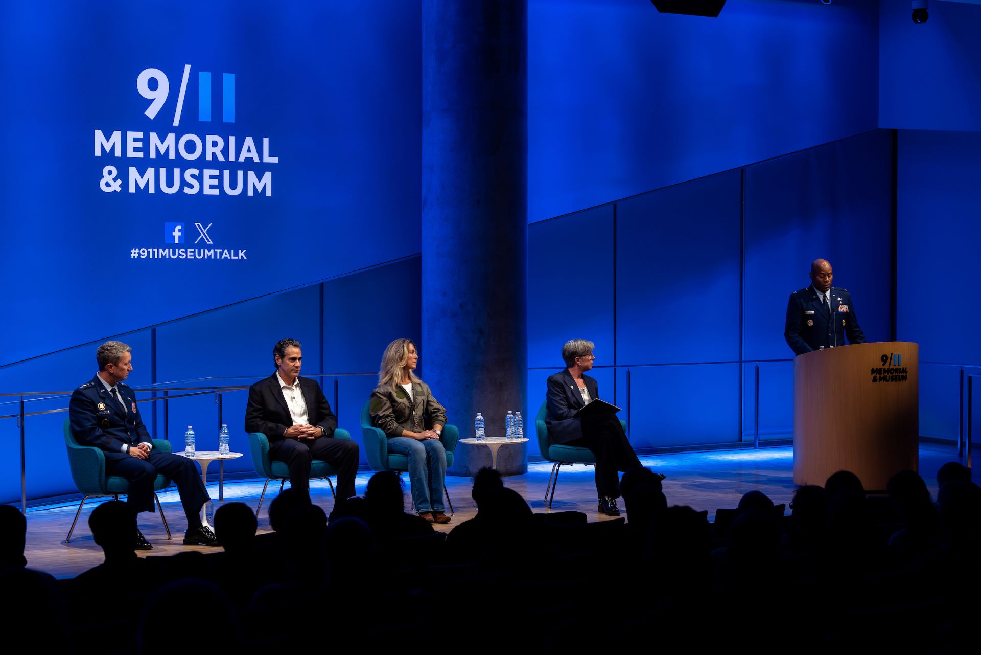 view of the blue background and all panelists being introduced, four seated, one standing person