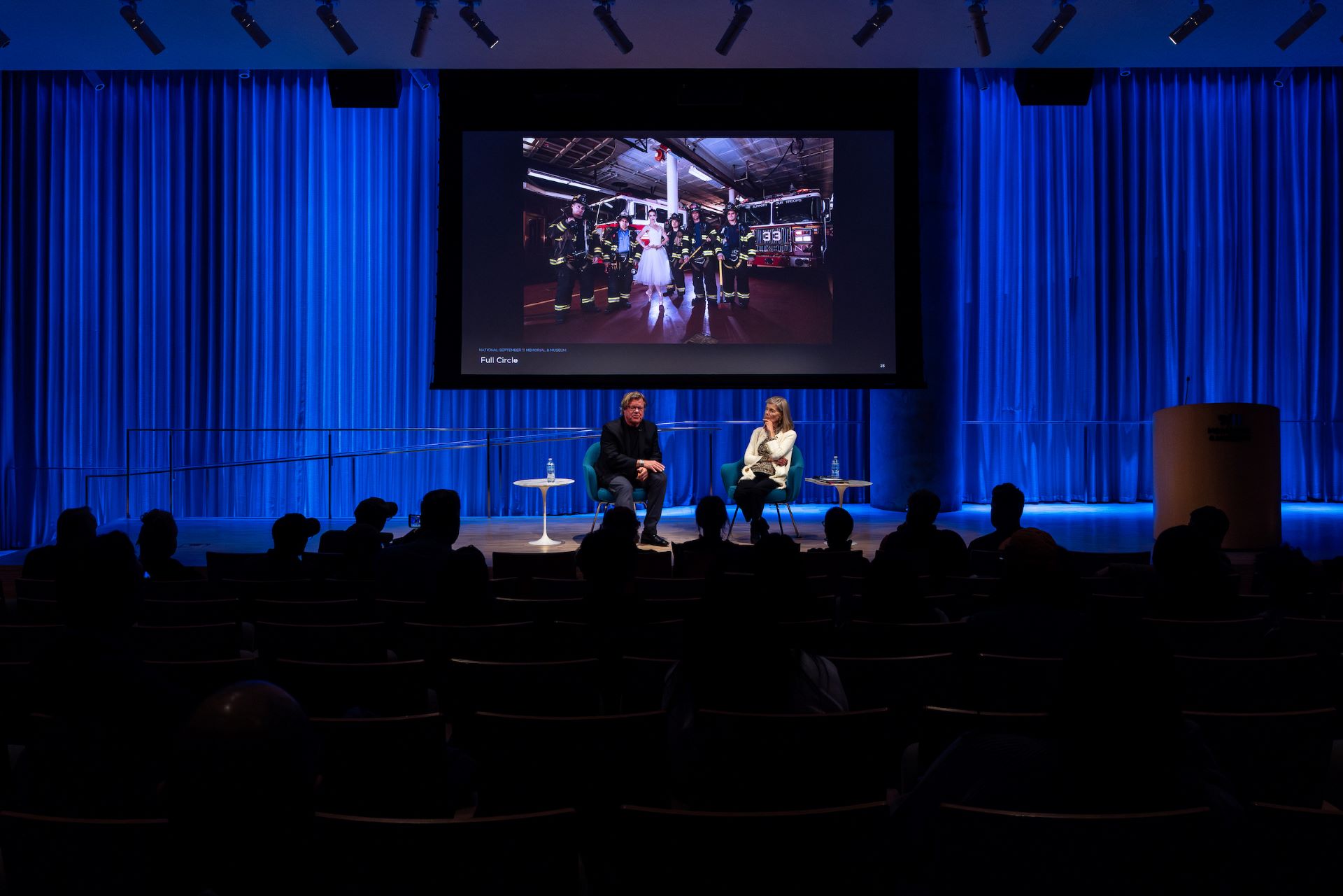 A wide lens shot of the audience from the back, featuring Joe McNally and Jan Ramirez on stage in front of a large screen featuring a slide