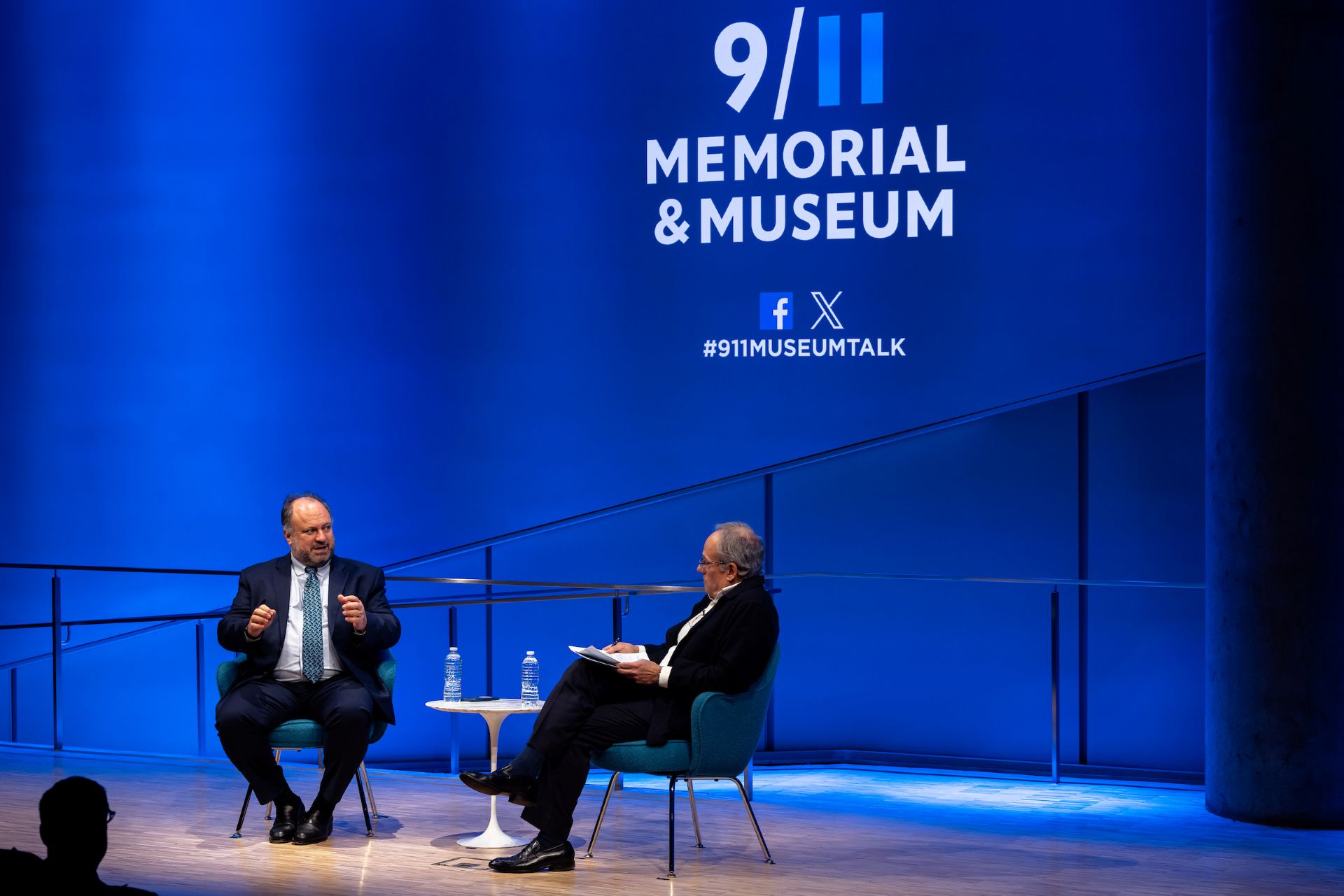 A wide shot of Bernie and Cliff speaking on the stage with the 9/11 logo visible