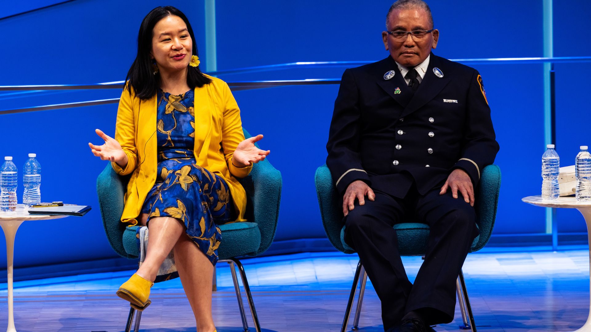 Two seated panelists on stage against a bright blue backdrop. 