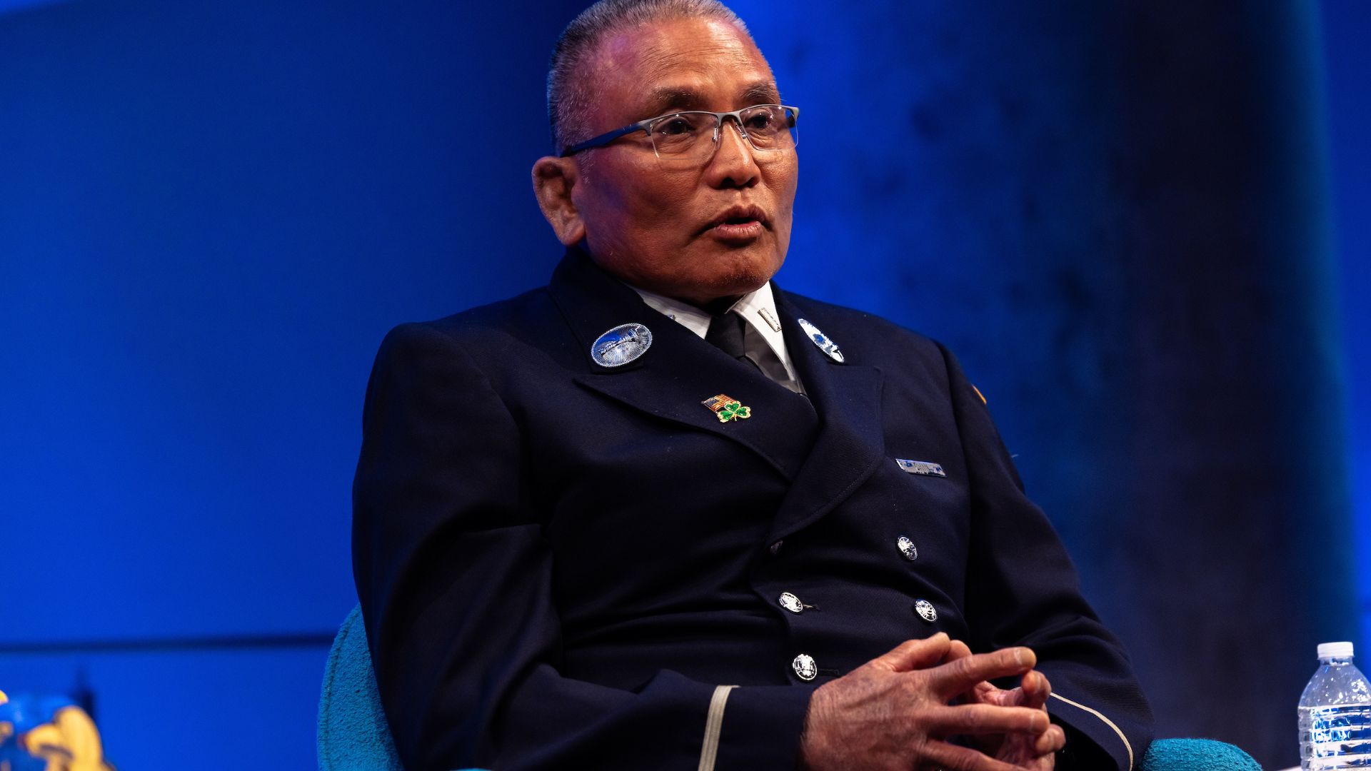 A panelist in an FDNY uniform sits with hands folded on stage against a bright blue backdrop. 