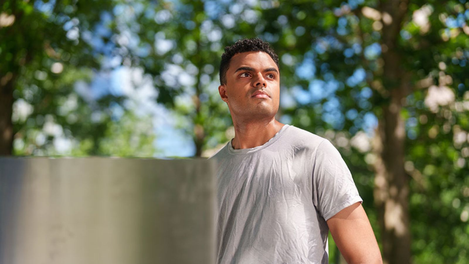 A man in a gray short-sleeve tee shirt, with short, dark, curly hair, looks into the distance with blurred trees behind him