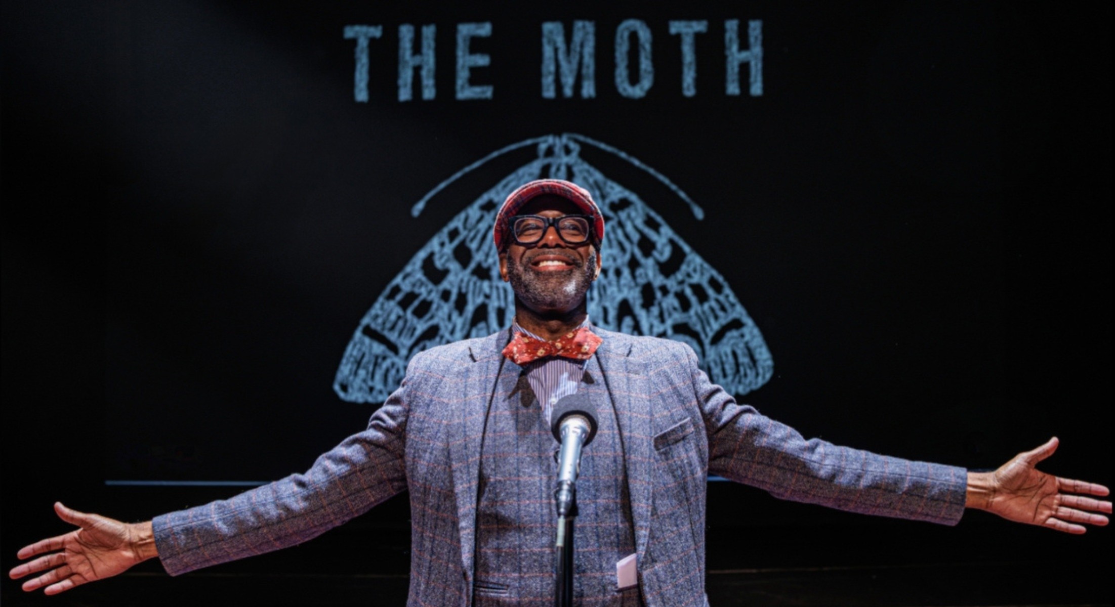 Jon Goode wearing a hat, arms wide, in front of the THE MOTH logo