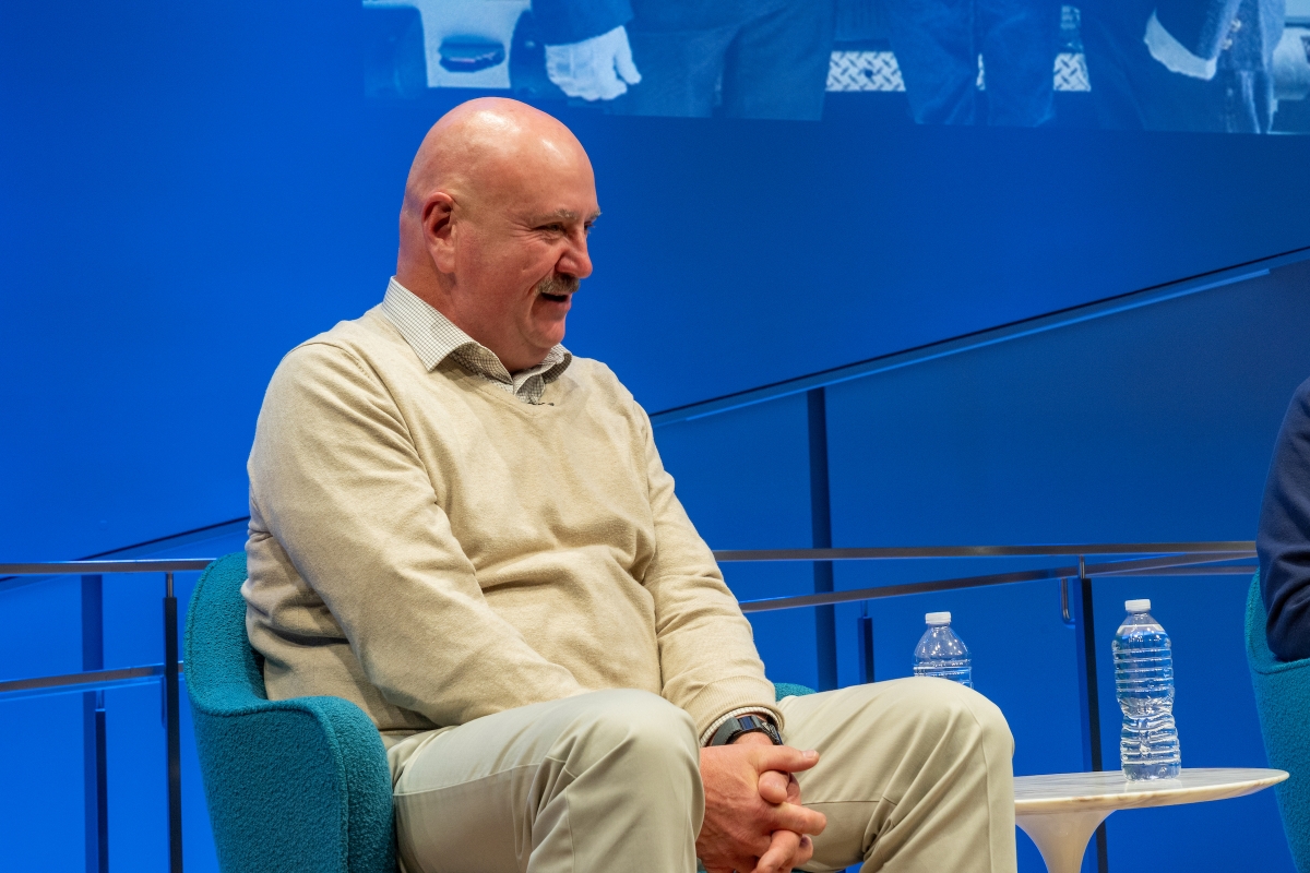 A man sits on stage in a chair with his hands folded.