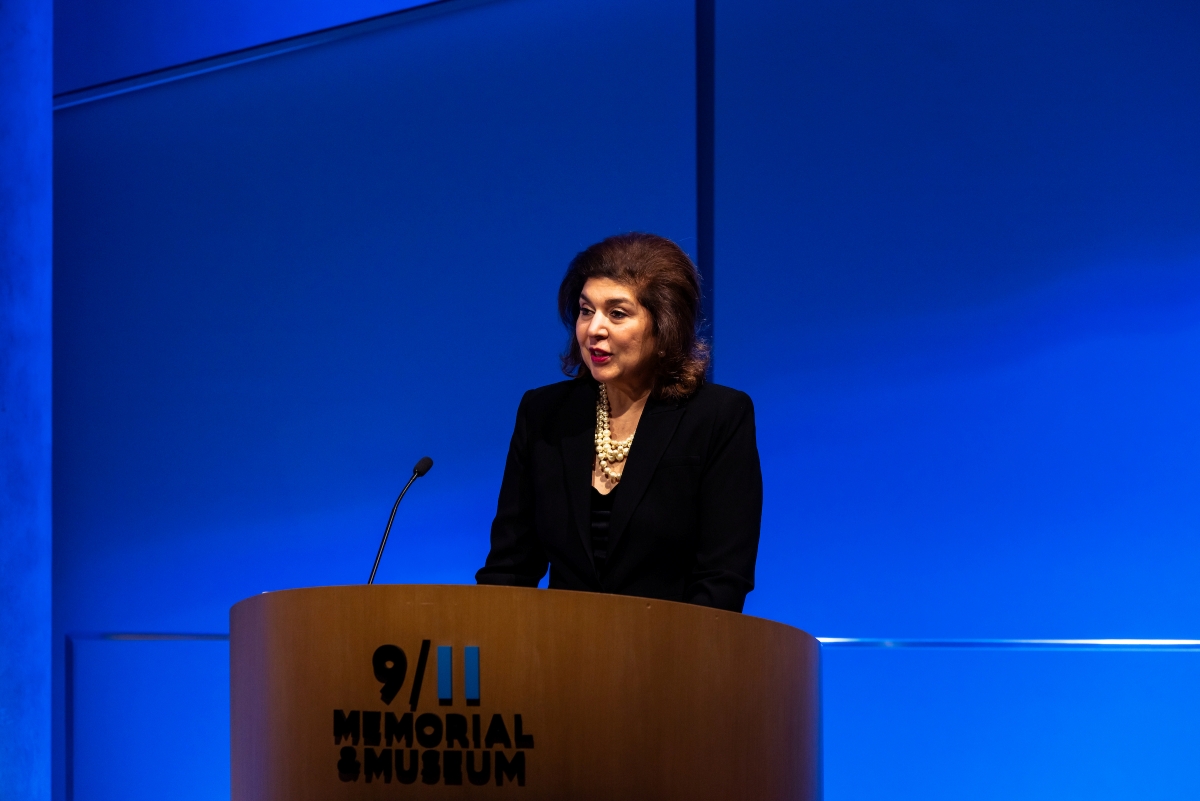 a woman in a dark blazer stands in front of a blue curtain behind the podium on stage