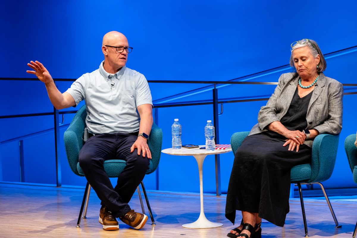 a man gestures on stage, across a small white table, a woman sits with her hands on her lap