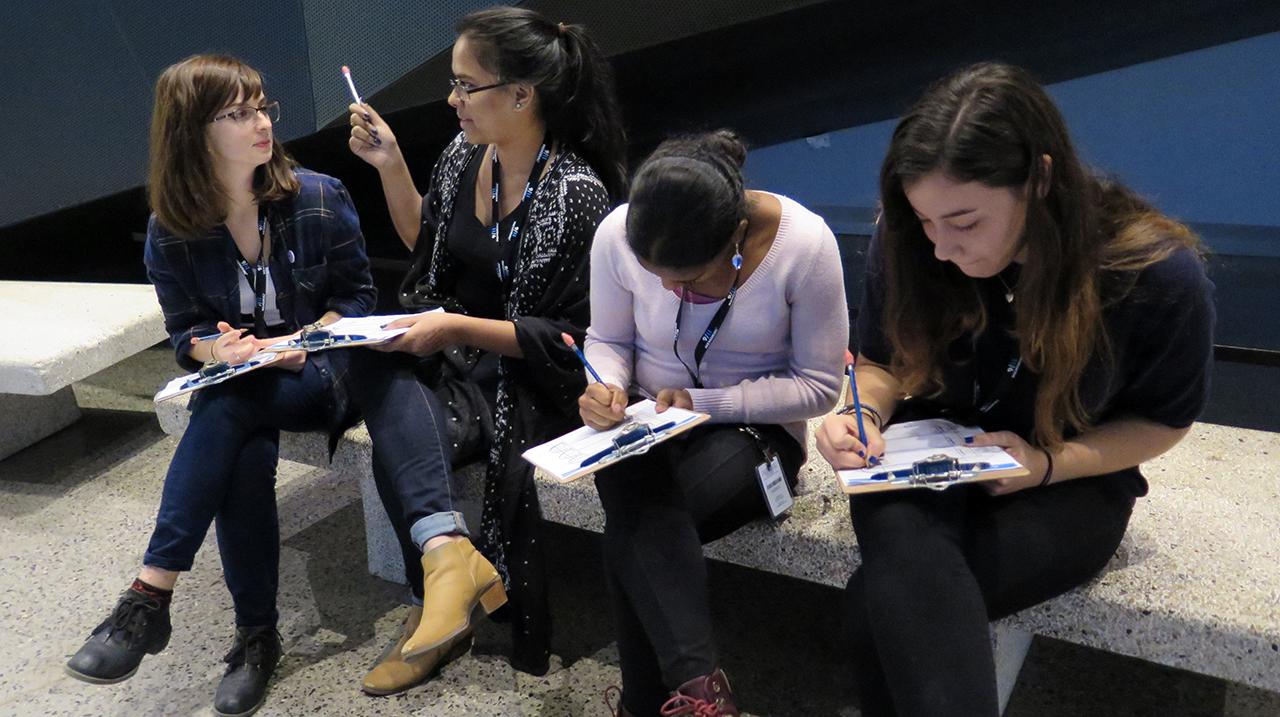 Four young women on a bench in the Museum. Two of them are writing on clipboards as the two others have a conversation.  