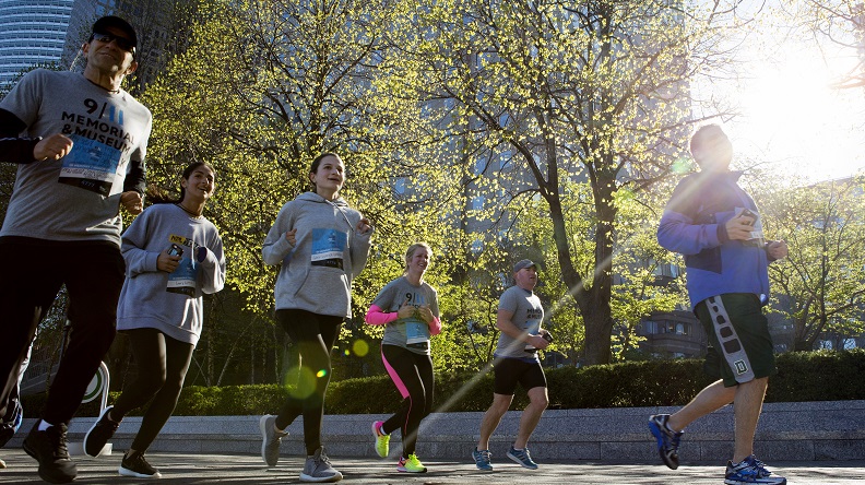 A half-dozen people in jogging clothes sprint down a street during the annual 5K Run/Walk. It’s morning and sunlight shines through the budding trees above them.