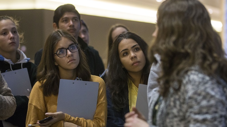 A group of students hold clipboards as they listen intently to a Museum guide in the foreground.
