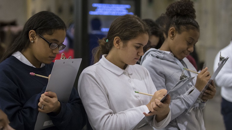 Three girls in school uniforms write on clipboards with their pencils. The Slurry Wall is seen behind them.