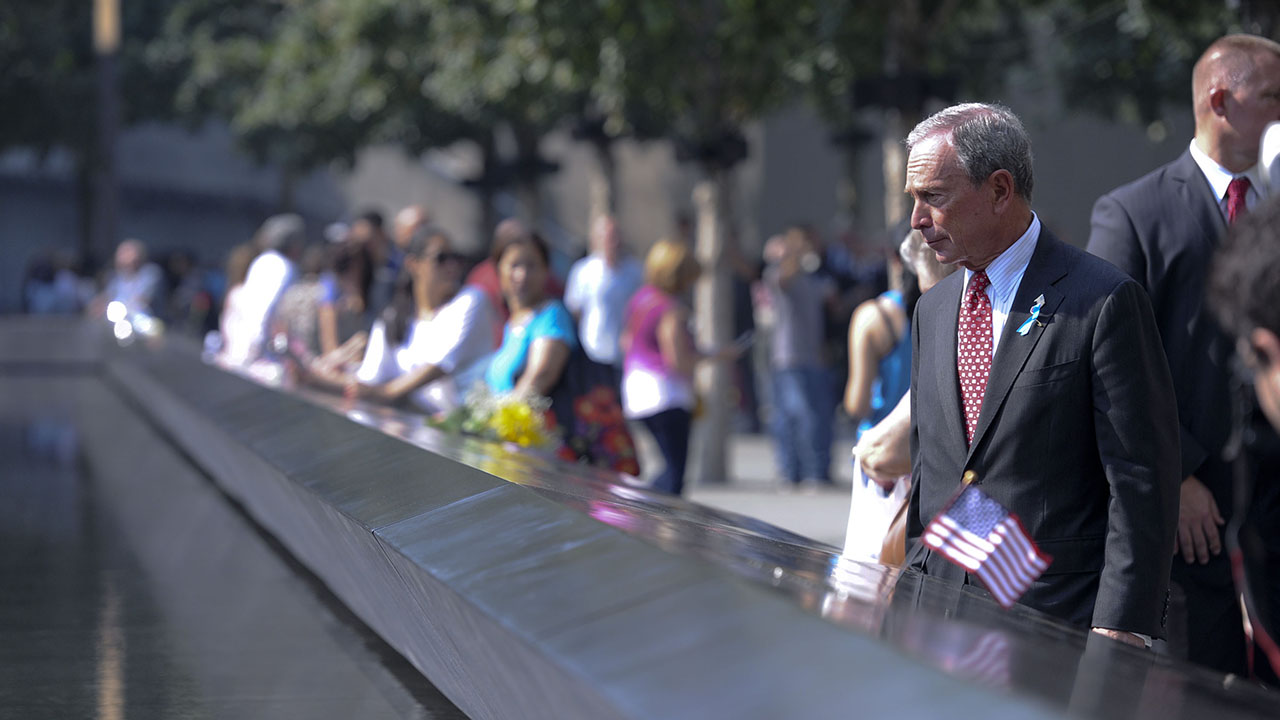 Michael Bloomberg, the chairman of the 9/11 Memorial & Museum, stands in a suit and tie beside a Memorial reflecting pool. In front of him, a bronze parapet runs along the border of the pool. A small American flag has been placed on the parapet. 