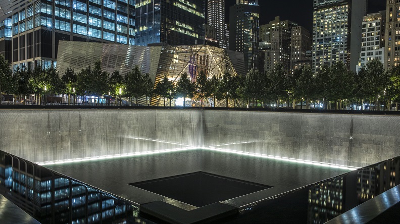 A reflecting pool of the Memorial is lit up at night. Illuminated water falls down the walls surrounding the expansive, square pool before disappearing down a square hole in its center. Lights from the Museum Pavilion and surroundings buildings enclose the Memorial. 