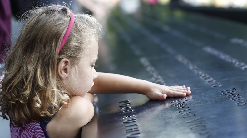 A young girl with blonde hair places her hand on a name etched in a bronze parapet at the Memorial.