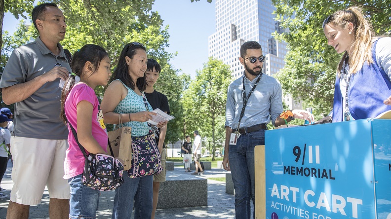 A man, woman, boy, and girl watch as two Museum employees display artwork at the Art Cart on the Memorial. It’s a warm day and sunshine falls on the trees and buildings in the background.