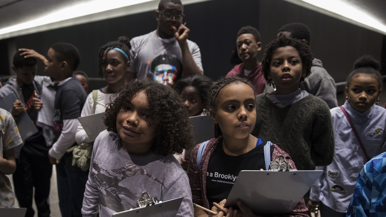 About a dozen children take part in a school program at the museum. The children are facing toward the photographer and holding clipboards.