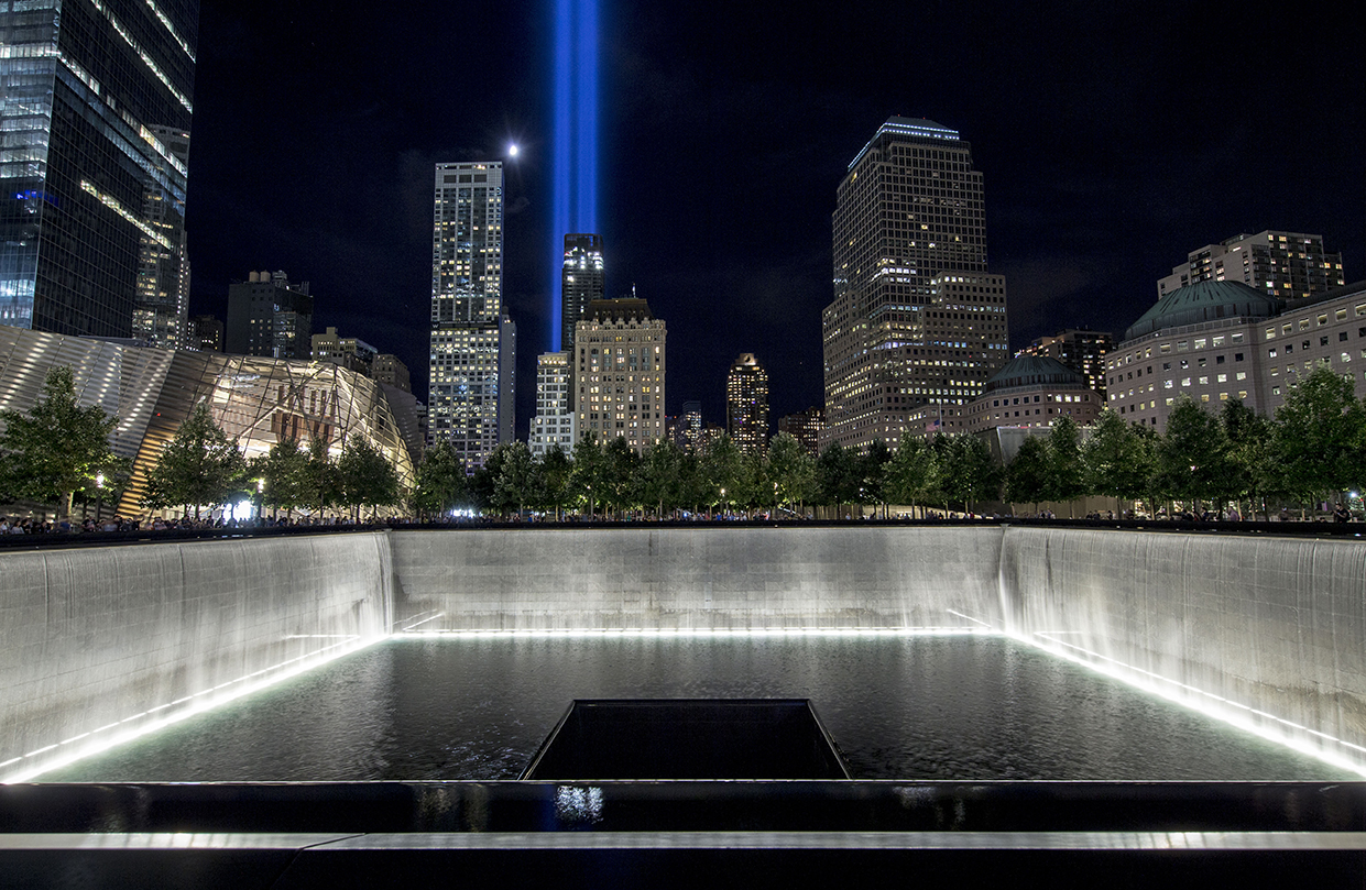 Water cascades down the illuminated walls of the North Tower reflecting pool on a warm night. The water pours down a square hole at the center of the pool. In the distance, a moon hangs over the city and the Tribute in Light shines above the buildings.