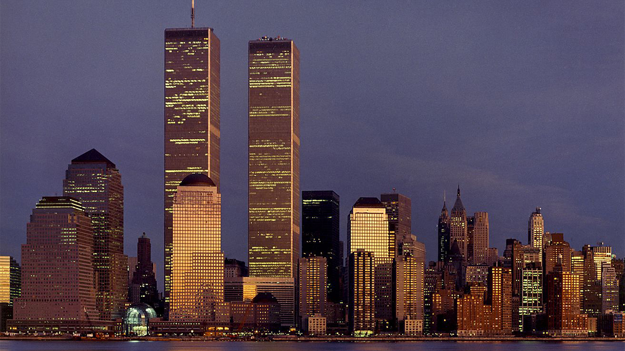 The Twin Towers dominate the skyline of lower Manhattan at dusk. An orange sunset reflects off of them and surrounding skyscrapers. The sky is a dark blue with a streak of clouds.