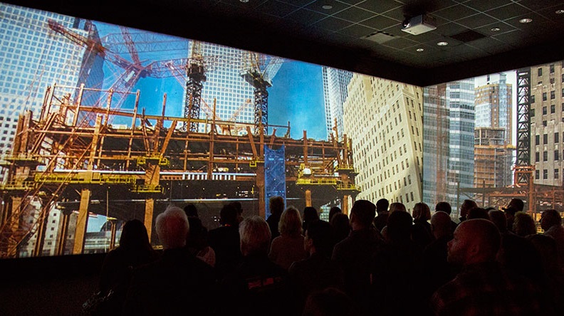 An audience of several dozen people watches a panoramic screen showing rebuilding at the World Trade Center site as part of the Rebirth at Ground Zero exhibition.