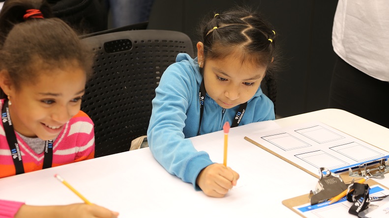 Two young girls sit at a white table and write on paper with pencils as they take part in a school program at the Museum.