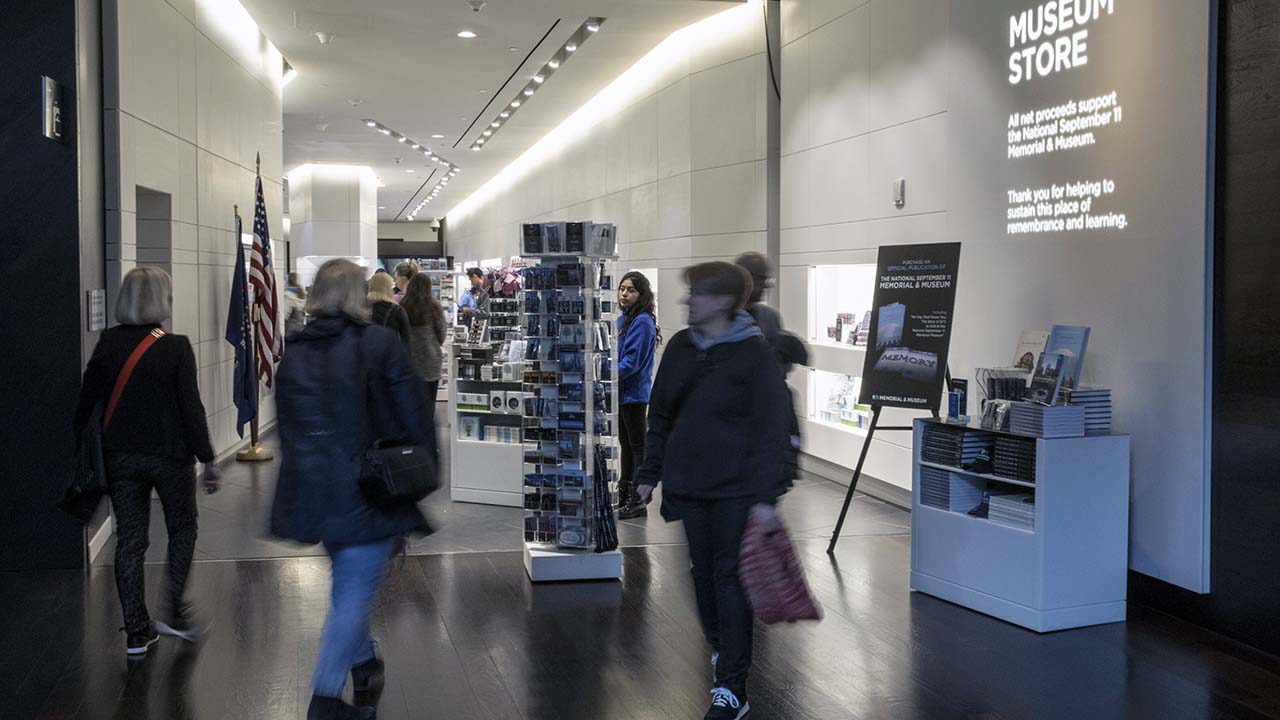 About ten people walk through the museum store. In the foreground, several visitors come and go from the shop. Further back, more people browse stands displaying postcards and souvenirs.