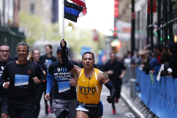 A man in a yellow NYPD Running Club shirt holds up a “thin blue line” flag and an American flag as he runs in the 9/11 Memorial & Museum 5K Run/Walk. Several other men are running beside him on a street in Manhattan. Onlookers watch from the sidewalk.