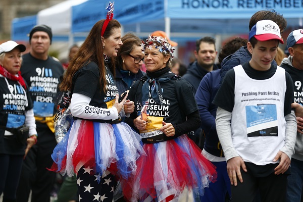 Two women wear patriotic outfits, including red, white, and blue tutus and American flag running pants, as they take part in the 9/11 Memorial & Museum 5K Run/Walk. Participants around them are also in running gear as they attend the event.