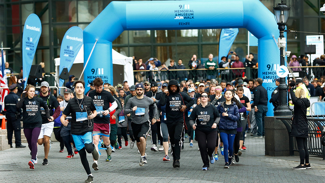 Dozens of participants pass through a blue, inflatable finish line arch at the 9/11 Memorial & Museum 5K Run/Walk. Spectators watch in the distance.