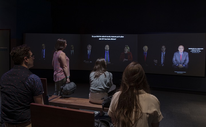 Four visitors watch a video screen that shows survivors recounting their memories of 9/11 in the Reflecting on 9/11 exhibition. Eight survivors can be seen in a panorama across the long video screen.