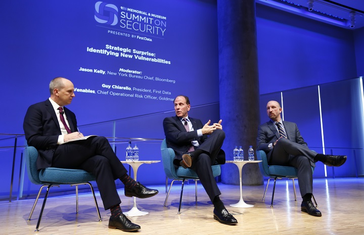 Three men in suits sit in chairs onstage at the Summit on Security. A projected outline of the event behind them says the event they’re taking part in is about “identifying new vulnerabilities.”