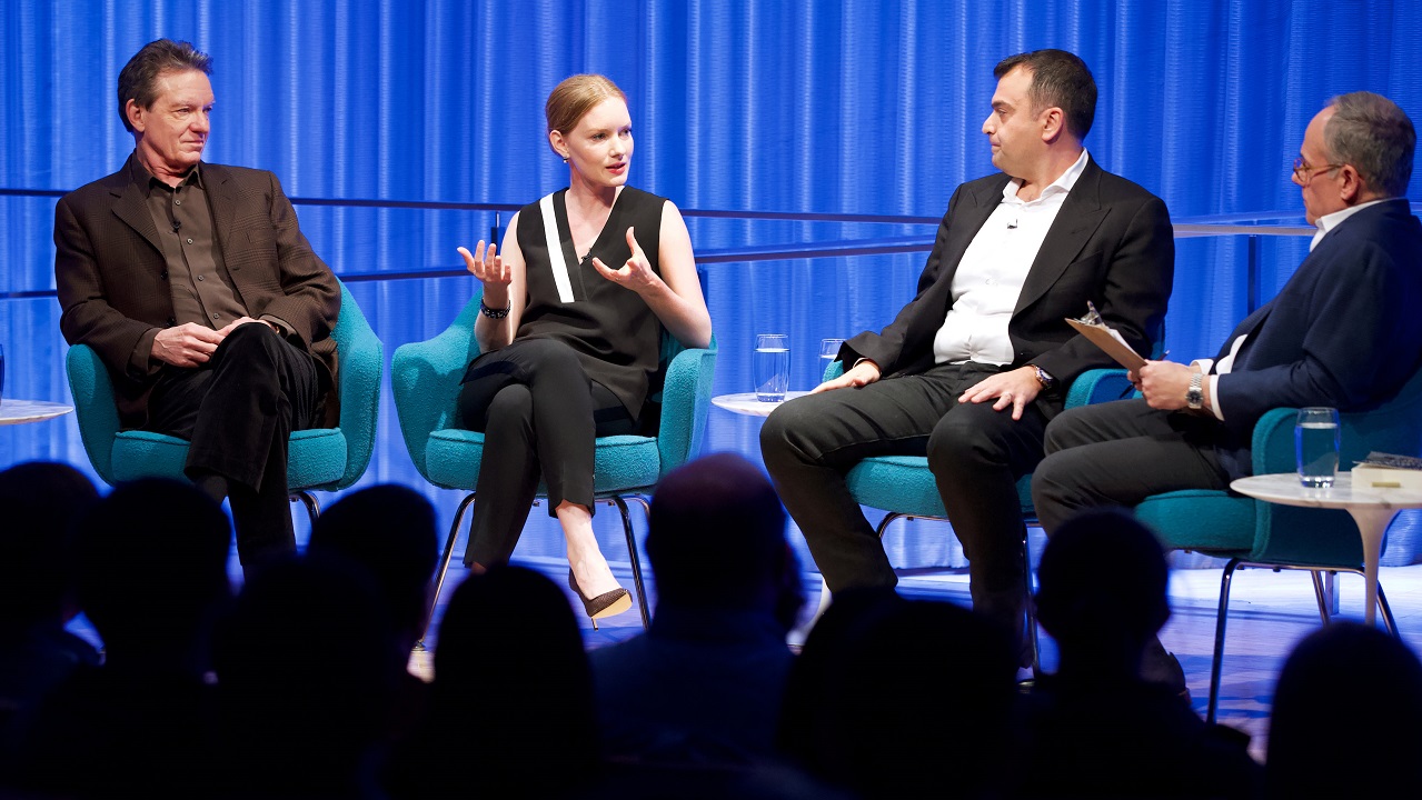 Three men and a woman take part in a moderated discussion on the auditorium stage at the Museum. The woman is speaking, second from the left, as the three men listen. The black silhouettes of audience members are in the foreground.