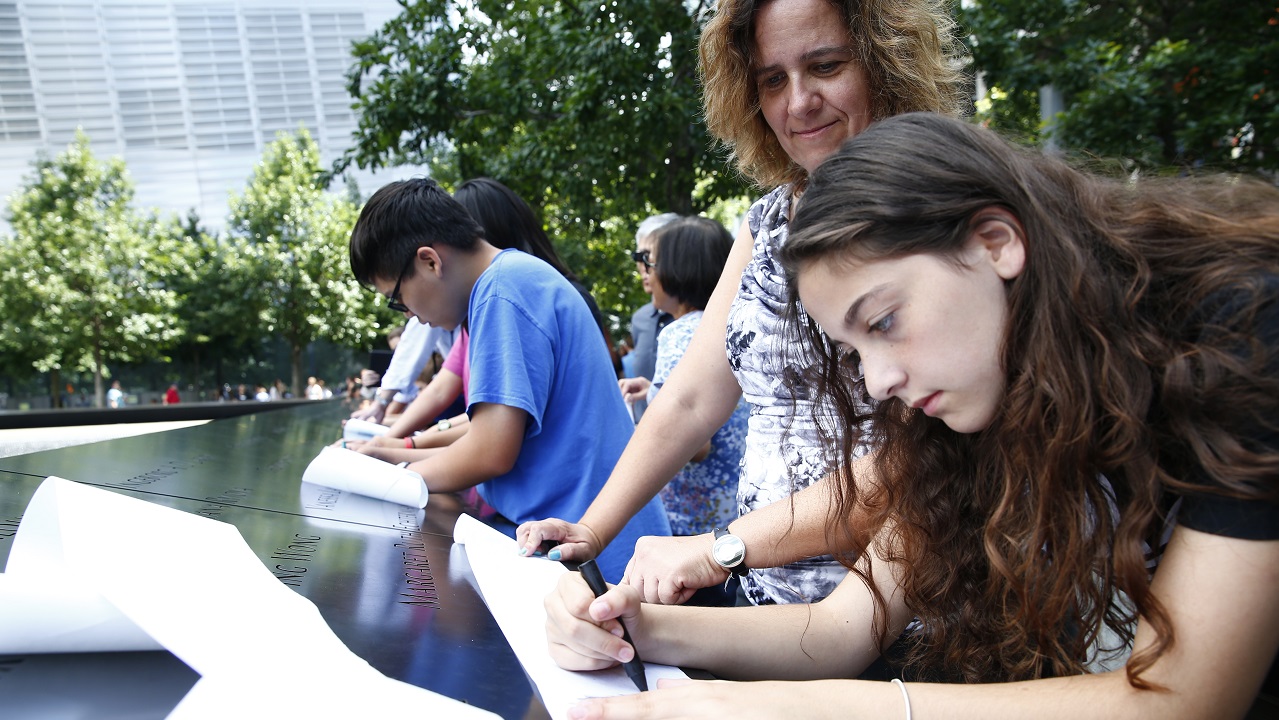 A teenage girl and a woman create a name impression at the Memorial by rubbing charcoal over a sheet of paper placed on an inscription. Other families beside them do the same along the bronze parapet inscribed with victims’ names.