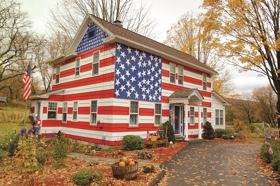 A house painted in the design of a large American flag is seen in a rural, autumn setting. The photograph is part of the exhibition Beyond Ground Zero: 9/11 and the American Landscape, Photographs by Jonathan C. Hyman. 