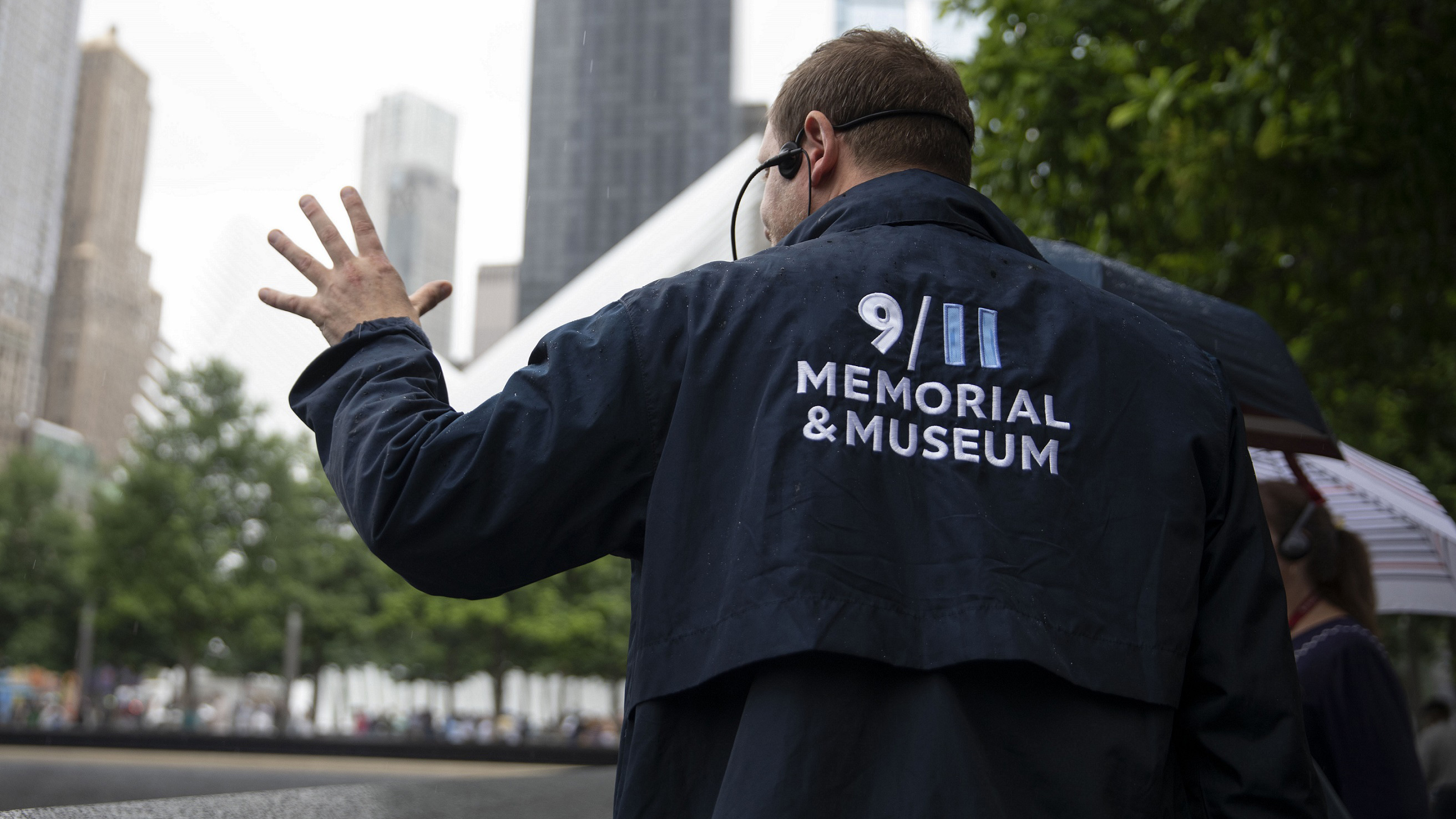 A tour guide in a blue 9/11 Memorial & Museum jacket faces away and motions with his left hand as he shows visitors a reflecting pool at the Memorial.