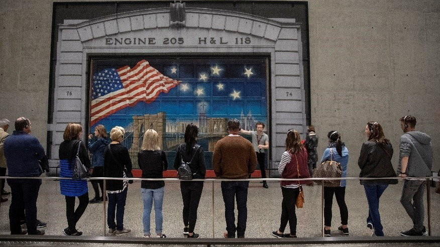 Ten visitors leaning on the railing, facing a young male tour guide explaining about a mural painted on the firehouse door behind him. The mural shows the skyline of lower Manhattan’s buildings along the river with a bridge spanning the river. A rippling large American flag and 8 bright stars dominate the evening sky.