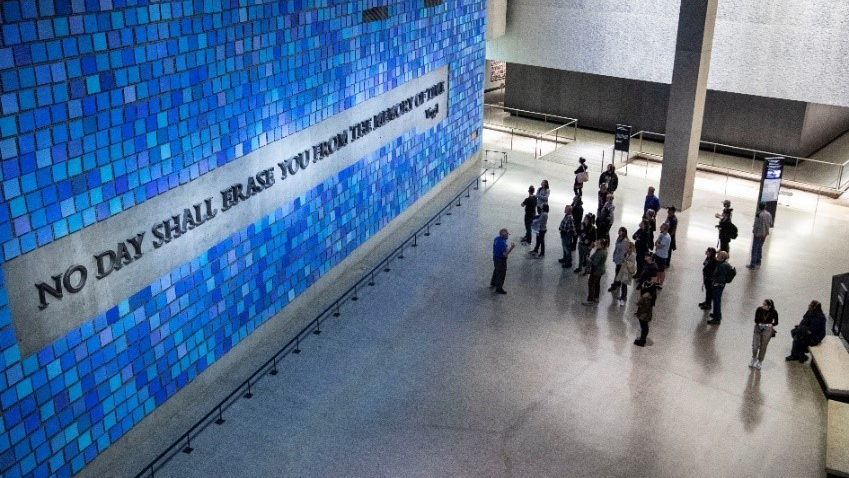An aerial view of a group visitors listening to a tour guide explaining about Virgil’s quote forged out of World Trade Center steel. The quote, “No day shall erase you from the memory of time,” is surrounded by a sea of individual square tiles in different shades of blue.