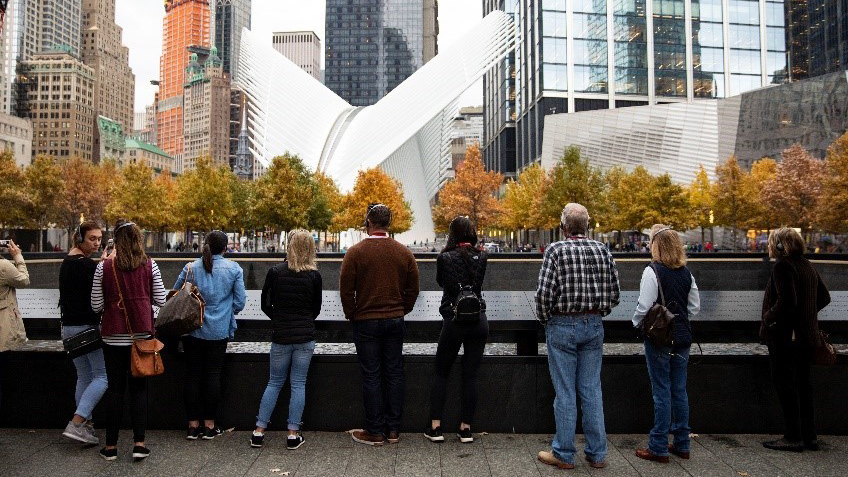 Visitors in autumn clothing stand beside the North Tower reflecting pool as they face towards the Oculus, a transportation hub with a white, winged design. The Oculus rises above fall-colored trees on the Memorial and is surrounded by the eclectic skyscrapers of lower Manhattan.