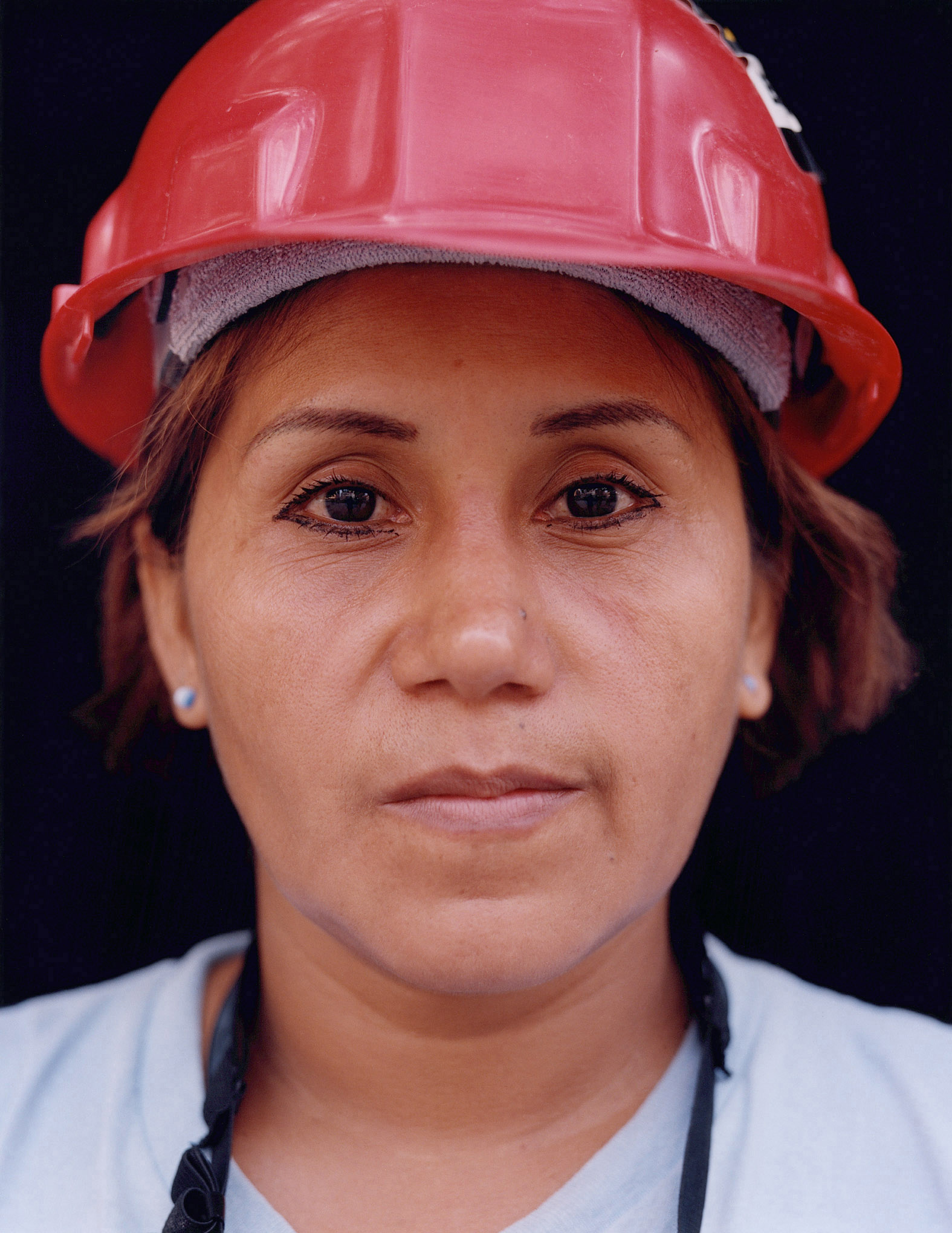 This close-cropped photograph shows a woman in a red hardhat, staring directly into the camera. 